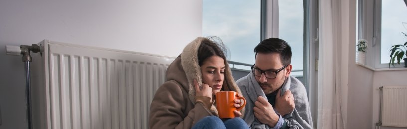 Man and woman sitting by broken radiator, cold and drinking a warm drink