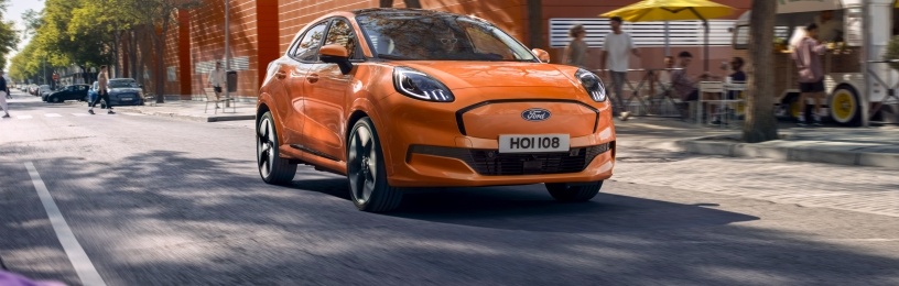 A bright orange EV drives past trees and cafés on a lively city street.