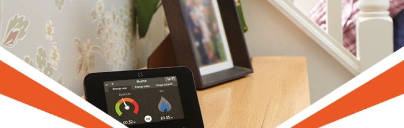 A smart meter on a mantelpiece in somebody's home. A photo frame with a blurred photo is placed behind it on the mantelpiece.