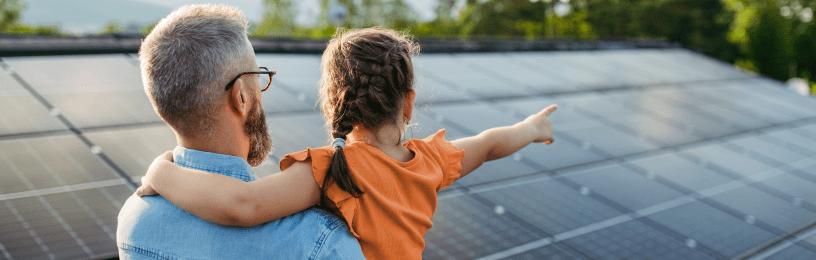 Man carrying girl in orange t shirt, pointing at solar panels 