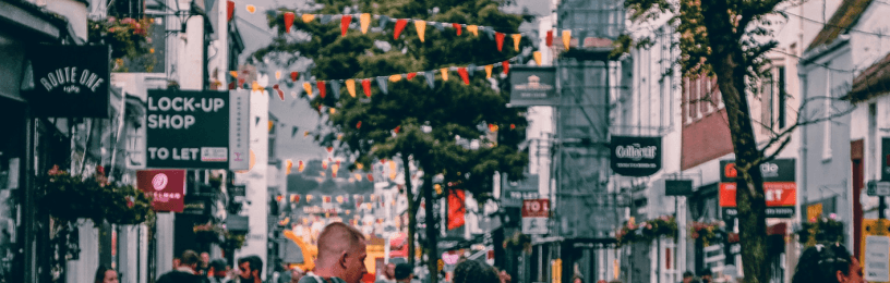 People walking along a busy high street lined with small shops, decorated with colourful bunting strung overhead.
