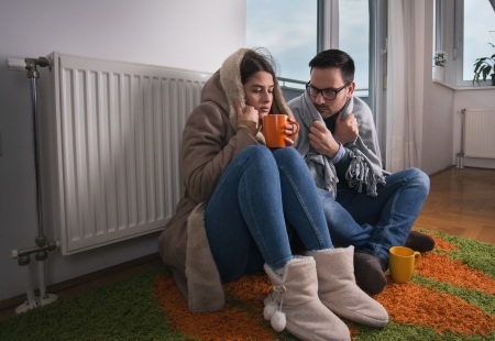 Man and woman sitting by broken radiator, cold and drinking a warm drink