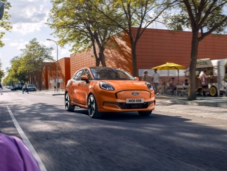 A bright orange EV drives past trees and cafés on a lively city street.