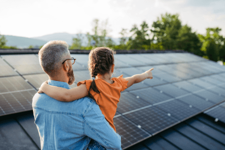 Man carrying girl in orange t shirt, pointing at solar panels 