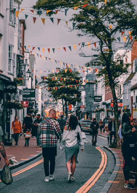 People walking along a busy high street lined with small shops, decorated with colourful bunting strung overhead.