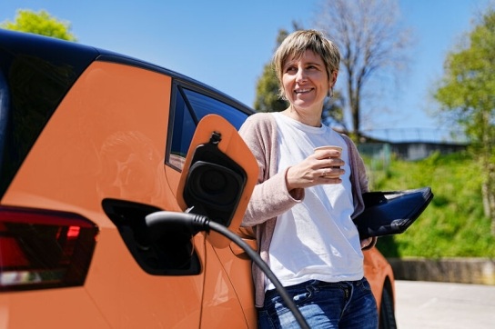 Woman in a white T shirt leaning on an orange electric vehicle while it charges
