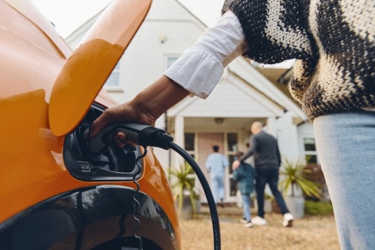 Person charging an orange EV with a house and people walking in to it, in the background