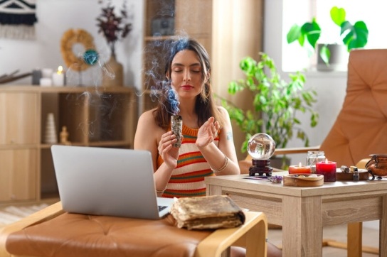 Woman sat in front of a laptop, burning sage with a crystal ball and candle next to her