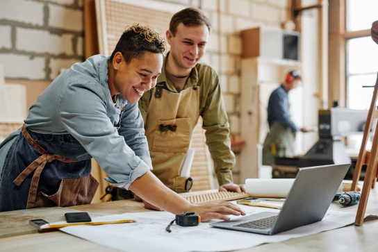 Two small business workers in a workshop lean over a laptop, reviewing plans on a workbench with tools and blueprints, while another person works in the background.