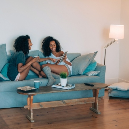 Two women relaxing on a sofa, laughing at something on a tablet